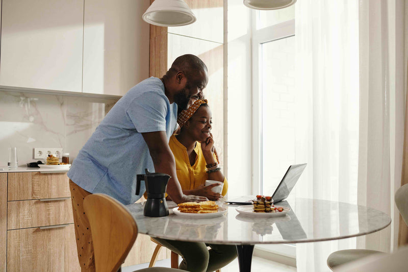 Two people at a kitchen table with a laptop, surrounded by food.