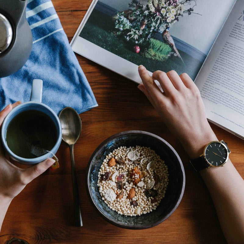 Person holding a mug with a bowl of healthy grains and nuts and magazine on a wooden table