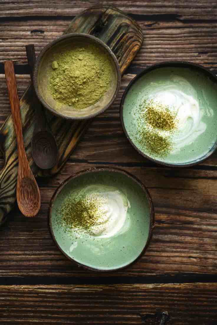 Three bowls of green matcha lattes and matcha powder on a wooden surface with a wooden spoon.