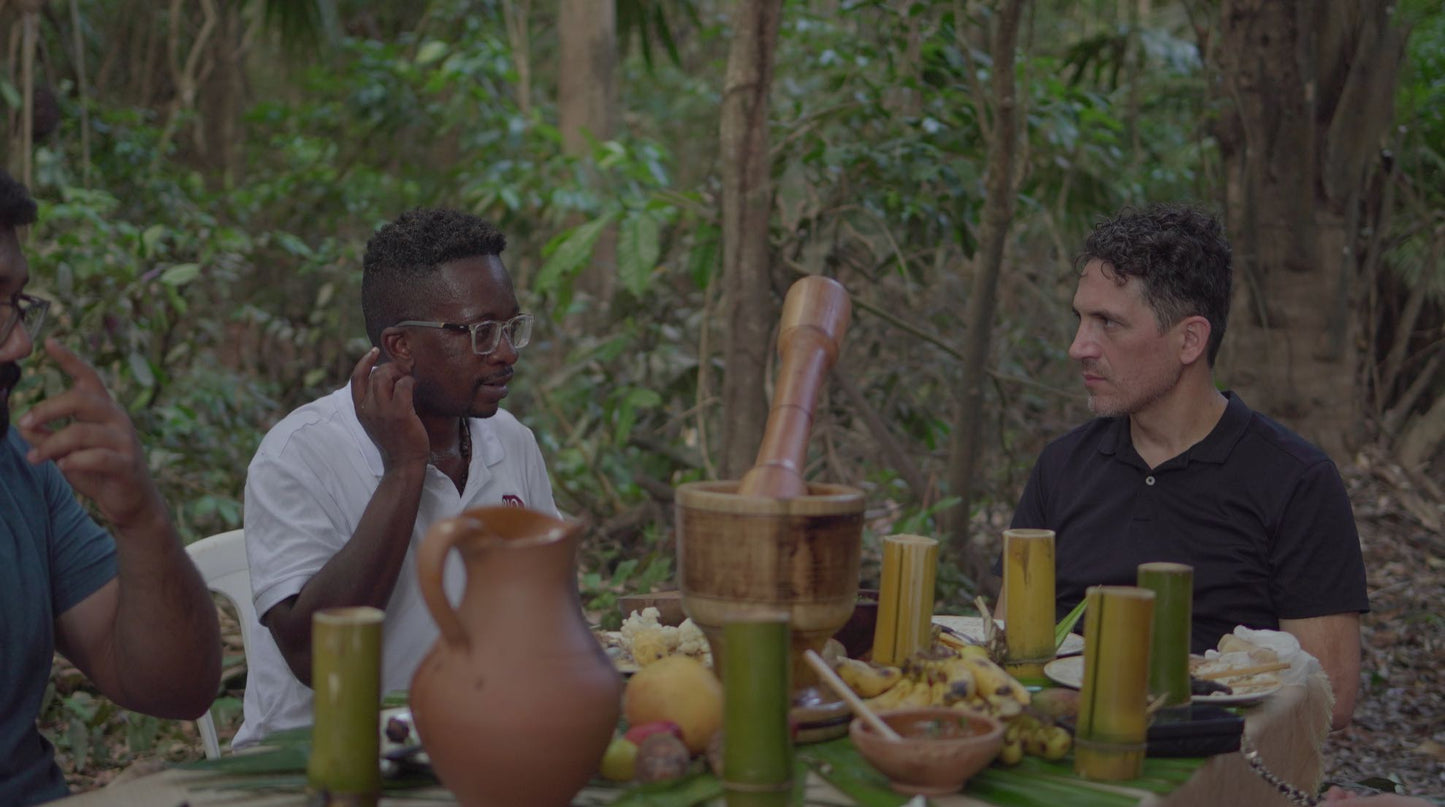 Three men sitting around a table with food and drinks in a forest setting